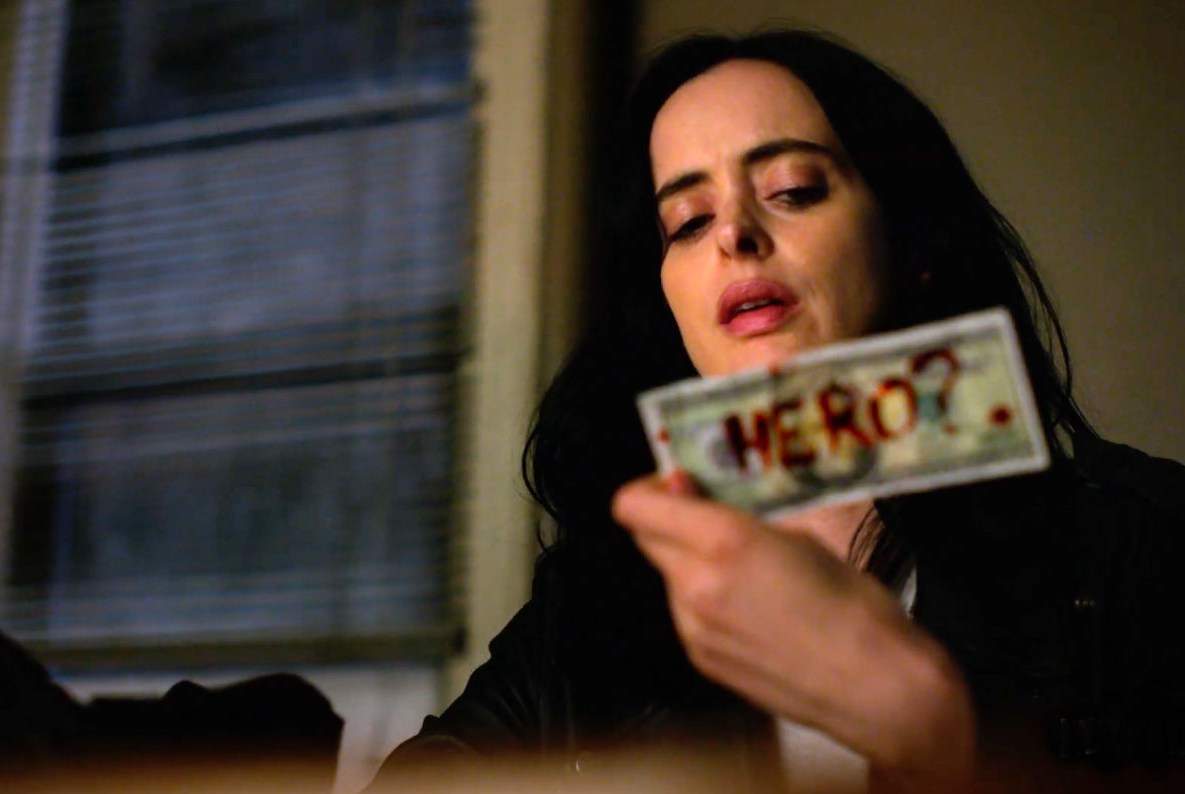 A screenshot of Jessica Jones from the Netflix series holding up a dollar bill onto which someone has scrawled the word “Hero?”