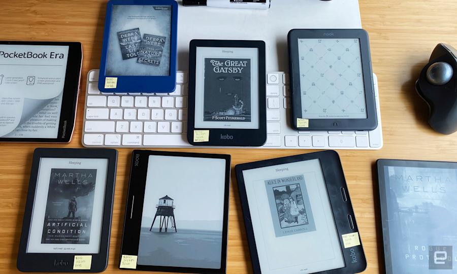An assortment of ereaders, including ones from Kobo, Kindle, Boox, Nook and PocketBook are arranged on a tan bamboo desk with a white keyboard and black mouse in the background.
