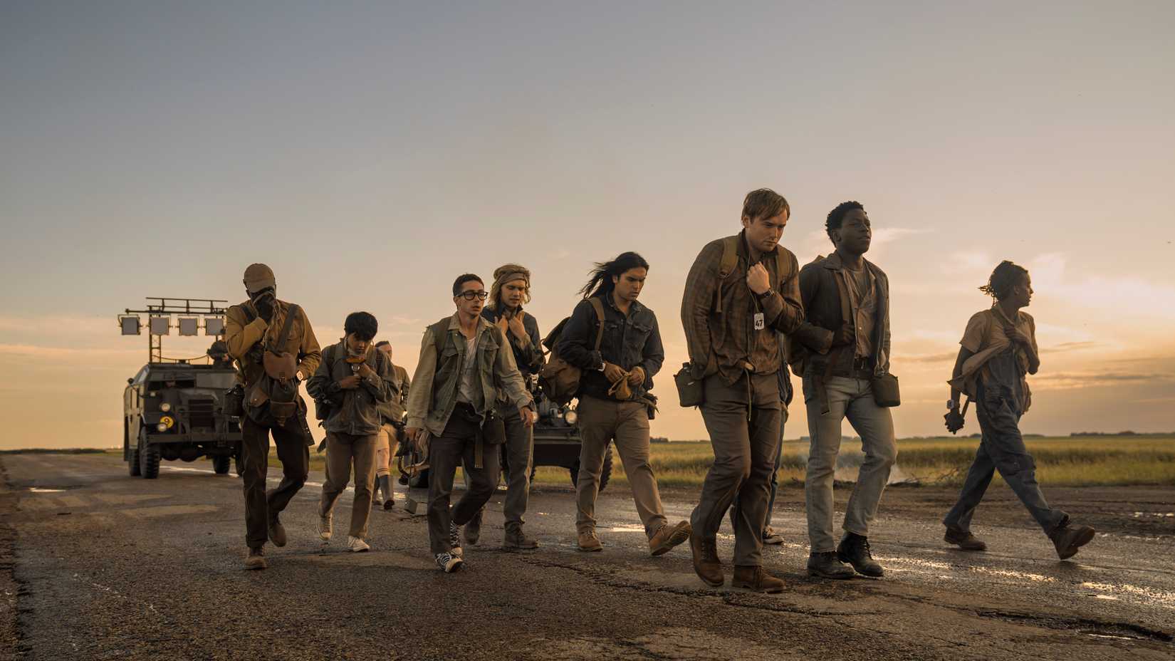 A group of teenage boys in earth-toned clothes walk along a road together near sundown in The Long Walk