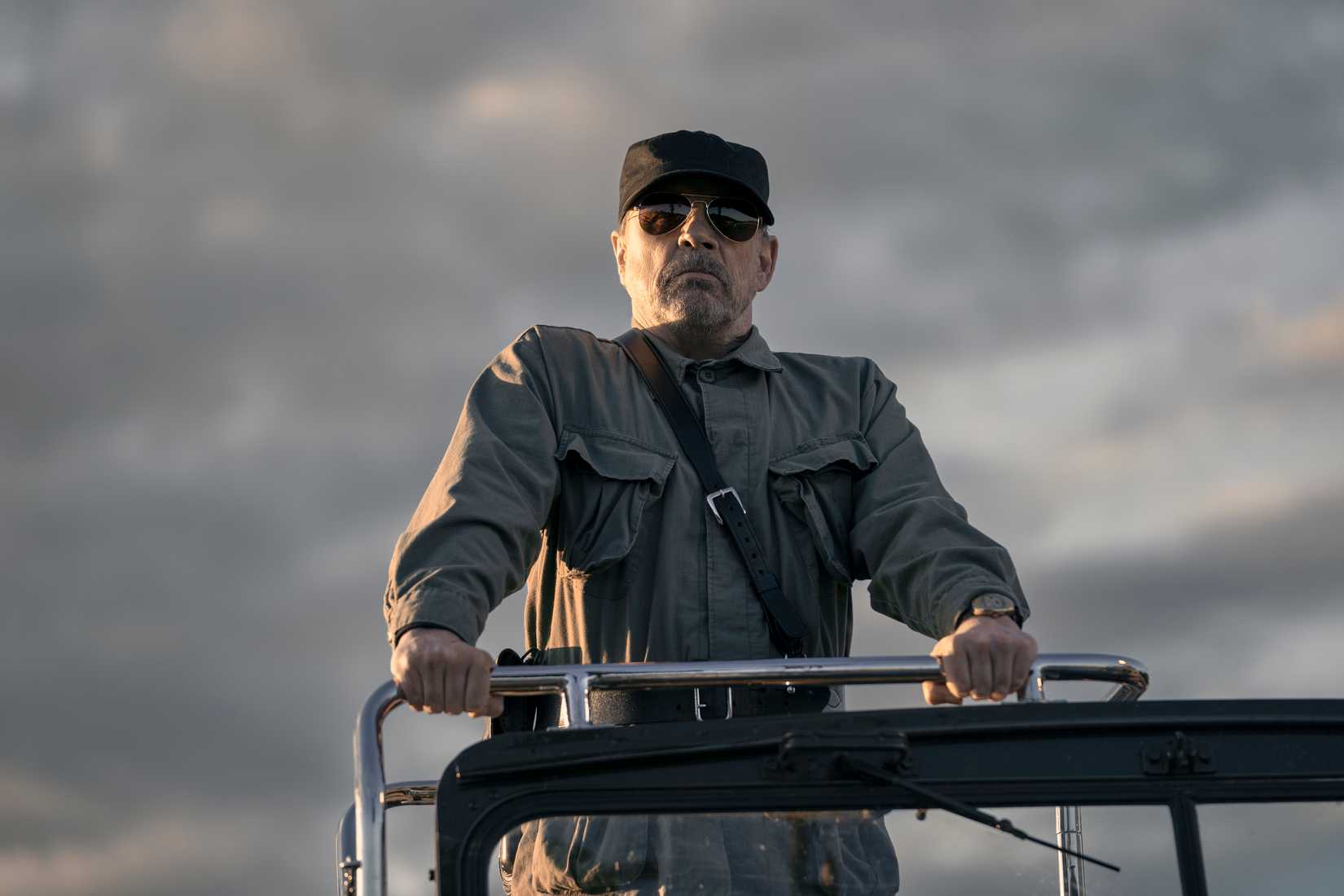 The Major (Mark Hamill), a man in a grey military uniform, black hat, and opaque sunglasses, stands in the back of a vehicle in The Long Walk