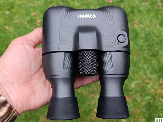 Overhead view of a hand holding the black Canon 8x20 Binoculars, more bulbous toward the front and tapering inward toward each eye-facing end.